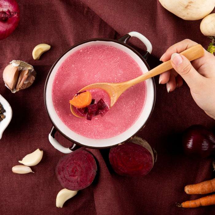 Pink soup being served with a wooden spoon, surrounded by ingredients on a red fabric background