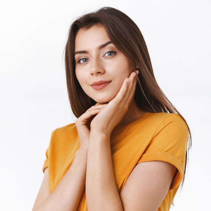 Woman in a mustard yellow shirt posing with one hand on her chin against a white background