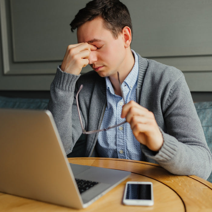 Man sitting at a table with a laptop and phone, holding his head in his hand.